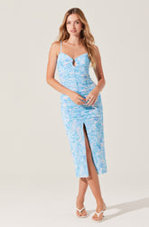 A woman is wearing a light blue, sleeveless, midi-length dress with thin spaghetti straps. The dress features a ruched bodice with a keyhole neckline and a front slit that extends to the knee. The fabric has a subtle, textured pattern in shades of blue. She is also wearing white, strappy high-heeled sandals.